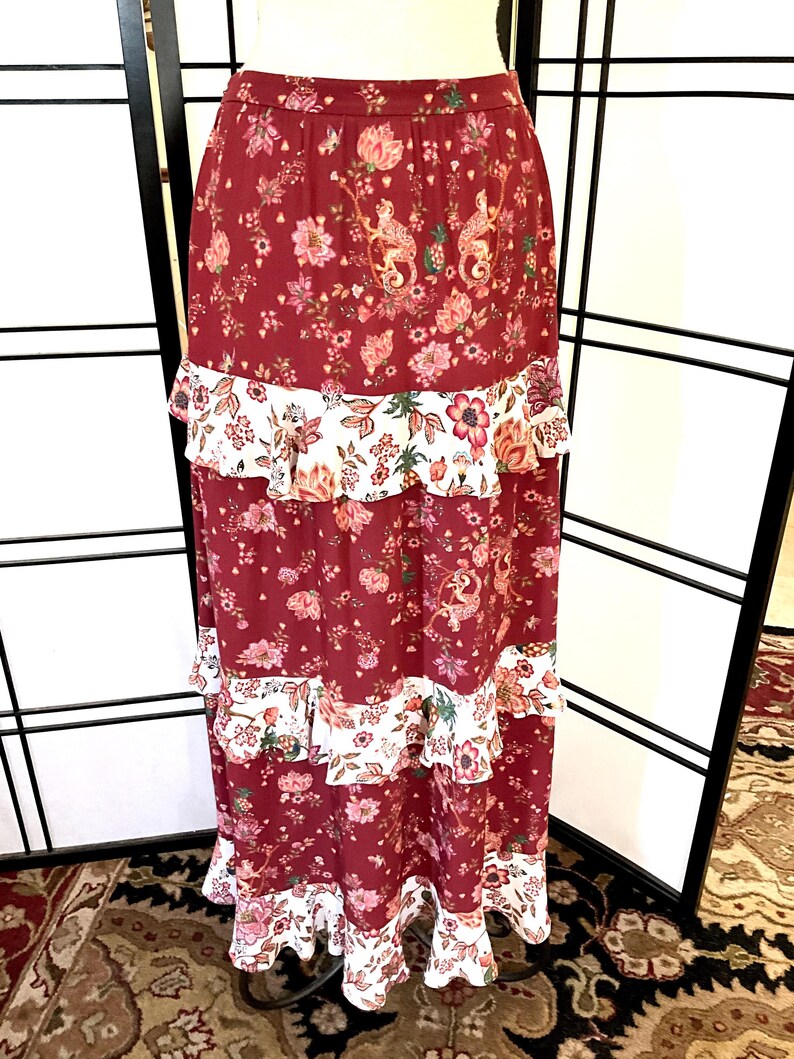 Pode incluir: Uma saia maxi longa, de cor bord&ocirc;, com estampa floral e v&aacute;rias camadas de acabamento branco com babados.