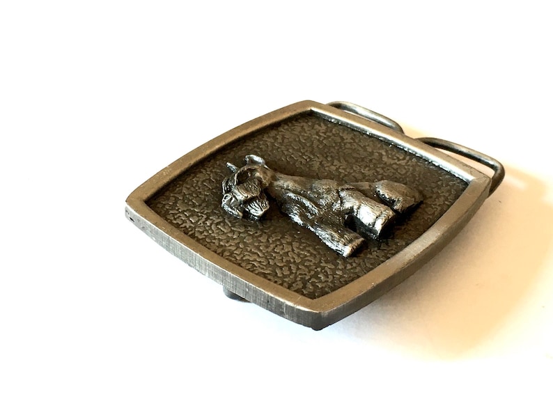 May include: A silver-toned metal belt buckle with a Schnauzer dog design. The dog is in a standing position on a textured, square background. The buckle has a rectangular frame with a single bar for the belt.