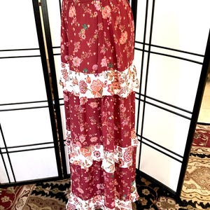 Pode incluir: Uma saia longa e em camadas com estampa floral em tons de vermelho, branco e rosa. A saia tem uma bainha com babados e uma cintura ajustada.