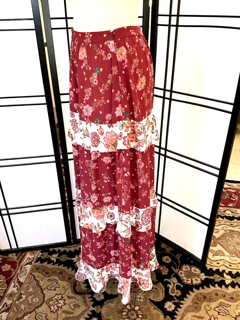 Pode incluir: Uma saia longa e em camadas com estampa floral em tons de vermelho, branco e rosa. A saia tem uma bainha com babados e uma cintura ajustada.