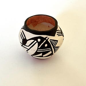 May include: Small, round ceramic pot with a black rim and interior. The exterior is white with black geometric designs and a reddish-brown interior. The pot is likely handmade and could be used for decorative purposes.