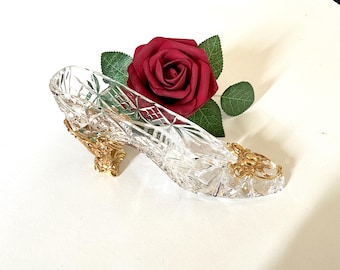 May include: A clear glass slipper with gold accents and a red rose on a white background.