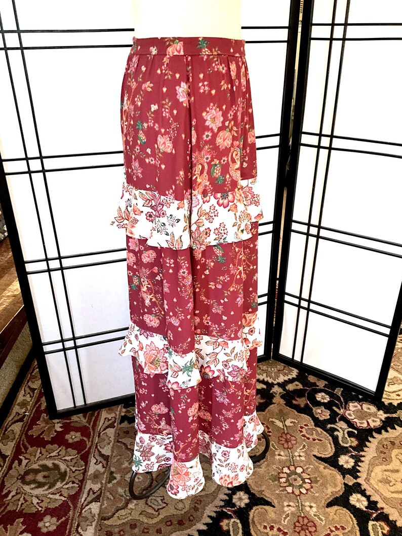 Pode incluir: Uma saia longa e em camadas com estampa floral. A saia &eacute; feita de um tecido macio e fluido e tem uma bainha com babados. A saia &eacute; de cor bord&ocirc; escura com detalhes florais brancos e rosados.