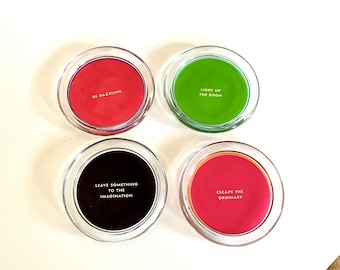 May include: Four round glass coasters with clear rims and solid coloured centres in red, green, black, and pink. Each coaster features white text: BE DAZZLING, LIGHT UP THE ROOM, LEAVE SOMETHING TO THE IMAGINATION, and ESCAPE THE ORDINARY.