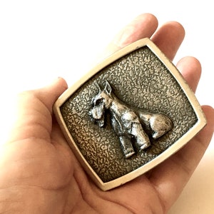 May include: A rectangular silver-coloured belt buckle with a detailed relief of a dog, possibly a Schnauzer, on a textured background. The dog is shown standing, with its characteristic beard and eyebrows visible.