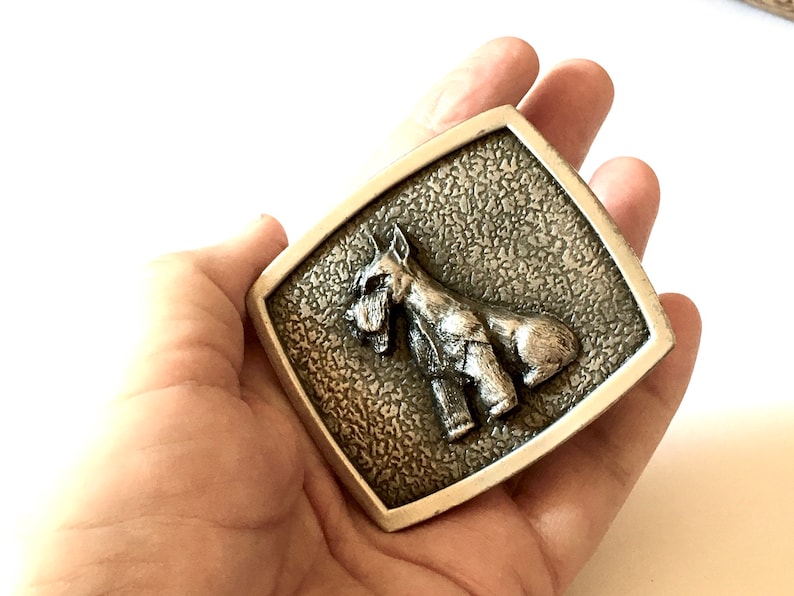 May include: A rectangular silver-coloured belt buckle with a detailed relief of a dog, possibly a Schnauzer, on a textured background. The dog is shown standing, with its characteristic beard and eyebrows visible.