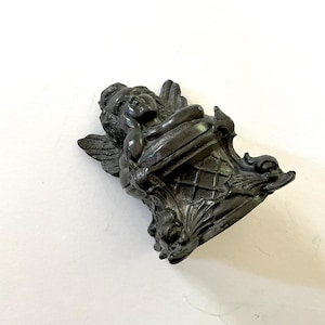 May include: A dark, antique-style cherub figurine. The cherub is reclining on a decorative urn-shaped base with ornate detailing. The figurine has detailed wings and is finished in a dark, possibly bronze, color.