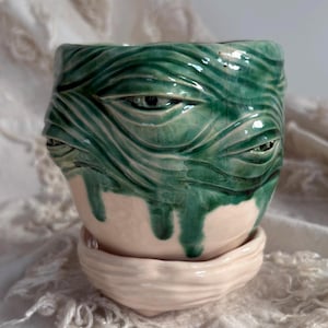 Puede incluir: Una maceta de cerámica con un esmalte verde brillante de acabado craquelado y detalles de ojos esculpidos. La parte superior tiene una superficie ondulada y texturizada con varios ojos, mientras que la base y el plato a juego son de color rosa pálido.