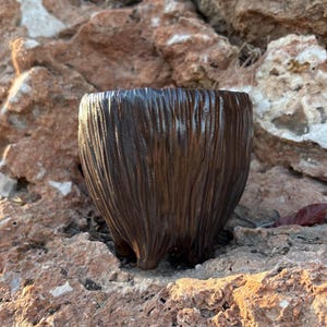 May include: A dark brown ceramic planter with a vertical, textured, bark-like finish. The vessel features a rounded, tapered body that rests on three short, integrated feet, creating a distinct silhouette with a deep, glossy glaze.