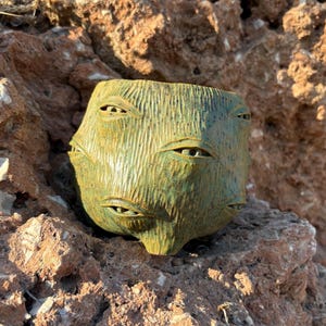 Puede incluir: Una taza de cerámica hecha a mano con un esmalte verde oliva texturizado y un acabado moteado. La superficie exterior presenta múltiples ojos esculpidos de aspecto realista incrustados en la arcilla, creando una apariencia de múltiples ojos en el recipiente.