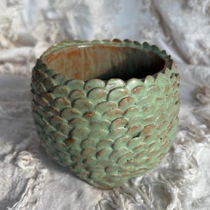 May include: A round ceramic planter featuring a textured exterior with overlapping green scale-like patterns and a smooth, warm brown interior. The vessel has a wide, open top and a rounded base, showcasing a handcrafted finish with visible colour variations.
