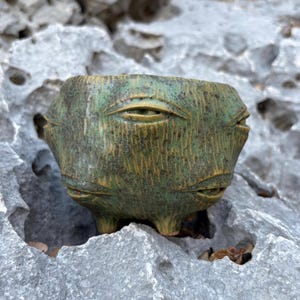 Könnte beinhalten: Ein kleiner, runder Pflanztopf aus grüner Keramik mit einer strukturierten, holzähnlichen Oberfläche. Das Gefäß ist mit mehreren skulptierten, geschlossenen Augen verziert, die in die Außenseite eingearbeitet sind, und steht auf drei kurzen Beinen.