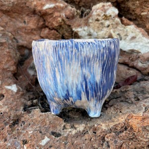 Puede incluir: Una pequeña maceta de cerámica con una superficie texturizada de costillas verticales. El exterior presenta un patrón de esmalte con vetas azul oscuro sobre una base de color blanco crema. El recipiente se apoya sobre tres pies cortos.