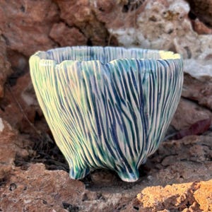 May include: A small ceramic planter with a textured, ribbed exterior featuring a glaze of blended teal, blue, and white tones. The vessel has a wide, slightly uneven rim and is supported by three short, integrated feet at the base.