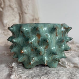 Op de afbeelding: Een keramische plantenpot met een glanzend blauwgroen glazuur en een getextureerd oppervlak met talloze uitstekende stekels. Het glazuur vertoont kleurvariaties met bruine en roestkleurige vlekken over de afgeronde, stekelige vorm.