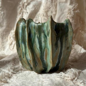 May include: A hand-built ceramic planter featuring a wavy, fluted exterior with a glossy glaze in shades of sage green, deep forest green, and earthy brown. The vessel has a distinct organic shape with vertical ridges and a textured finish.