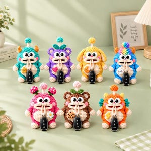 May include: Seven colorful, crocheted monster-shaped key covers, each featuring large white eyes, a wide mouth, and a small pom-pom on top. Each creature holds a black car key fob and displays a unique embroidered patch like a star, heart, or moon.