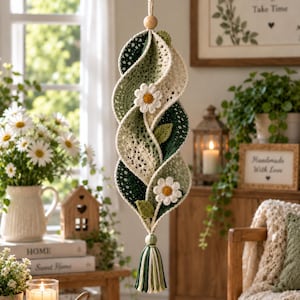 May include: A hanging crochet spiral ornament featuring alternating shades of cream, sage green, and dark forest green yarn. The structure is adorned with two white daisy appliques with yellow centers and small green leaves, finished with a tassel.