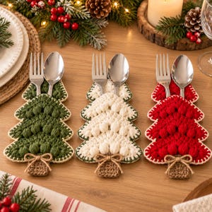 Könnte beinhalten: Drei gehäkelte Bestecktaschen in Form von Weihnachtsbäumen in Dunkelgrün, Creme und Rot. Jede Tasche hat ein strukturiertes Noppenmuster, einen braunen rechteckigen Fuß mit einer Bindfaden-Schleife und enthält jeweils eine silberne Gabel und einen Löffel.