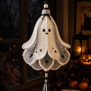 May include: A hanging crochet ghost ornament featuring a white and grey layered bell-shaped body with scalloped edges. The ghost has two black circular eyes and a small black smile, accented with tiny black star motifs and a black and white tassel.