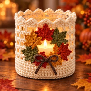 May include: A cylindrical glass candle holder encased in a cream-colored crocheted sleeve with a scalloped top edge. The sleeve features a decorative arrangement of crocheted autumn leaves in red, orange, and green, accented by a brown yarn bow.