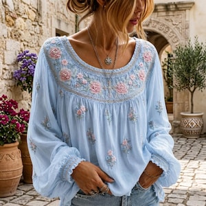 Op de afbeelding: Een lichtblauwe blouse met lange mouwen en een ronde halslijn met gedetailleerd roze en wit bloemenborduursel. Het kledingstuk heeft geplooide mouwboorden, een losse pasvorm en verspreid bloemenborduursel op de voorzijde en de mouwen.