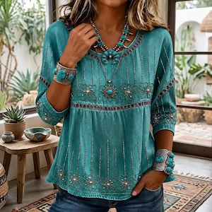 May include: A teal long-sleeve top featuring intricate silver embroidery, floral patterns, and vertical line details. The garment is styled with a large turquoise stone necklace and two matching wide silver cuff bracelets with turquoise stones.