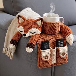May include: A crochet fox-shaped sofa armrest organizer in orange, white, and dark brown. It features a sleeping fox head, a matching cup holder with a white mug, and two pockets with wooden buttons containing black remote controls.