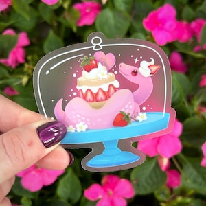 Op de afbeelding: Een roze en witte taart met aardbeien en een roze slang eromheen gewikkeld. De taart zit in een doorzichtige koepel met een witte basis. De achtergrond is een roze bloem.