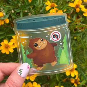 May include: A clear glass jar with a brown furry creature inside. The creature has a big smile and is standing on its hind legs. The jar has a green lid and a no photography sign on the front.