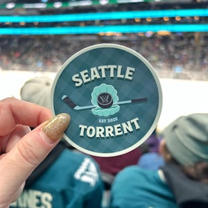 Puede incluir: Una pegatina redonda con las palabras "SEATTLE TORRENT" en blanco, verde azulado y negro. La pegatina presenta un palo de hockey, una concha y el texto "EST 2025". La pegatina es verde azulado con un patrón de diamantes.