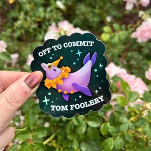 May include: A die-cut sticker with a dark green background featuring a purple seal wearing a yellow party hat and a ruffled orange collar. The text reads OFF TO COMMIT TOM FOOLERY in white capital letters, surrounded by small light blue stars.