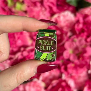 May include: A green and gold enamel pin in the shape of a pickle jar. The jar has a brown label with the words "PICKLE SLUT" in white, with hearts on either side. The pin is held in a person's hand.