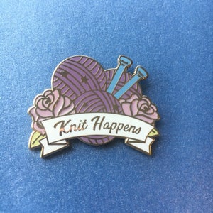 Può includere: Uno spillo in smalto viola e bianco con un gomitolo di lana, ferri da maglia e due rose rosa. Lo striscione recita "Knit Happens".