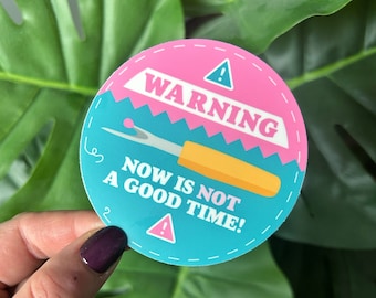 May include: A round sticker with a pink and blue design. It features a warning sign with the text "WARNING NOW IS NOT A GOOD TIME!" and a yellow sewing needle.