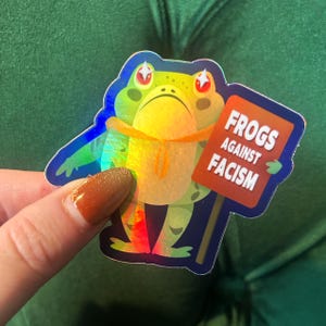 May include: A holographic sticker featuring a cartoon frog holding a sign that reads "Frogs Against Facism." The frog is green, yellow, and blue with red accents. The sign is orange with white text.