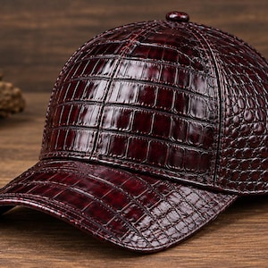Puede incluir: Una gorra de béisbol de color burdeos intenso con un acabado brillante y texturizado que imita la apariencia de la piel de cocodrilo. La gorra tiene una corona estructurada, una visera curva y un pequeño botón a juego en la parte superior.