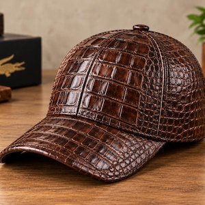 Puede incluir: Una gorra de béisbol de color marrón oscuro con un patrón de piel de cocodrilo brillante y texturizado en toda la superficie, incluyendo la visera curva y la corona de seis paneles con un botón a juego en la parte superior.
