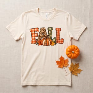 May include: A cream-colored short-sleeve t-shirt featuring the word FALL in large, patterned block letters. The letters include orange plaid, leopard print, and green floral designs. A central orange pumpkin graphic sits below the letters.