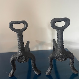 May include: A pair of black cast iron andirons featuring a textured, rugged surface. Each piece has a vertical post with a rounded, loop-shaped handle at the top and three curved legs extending outward to provide a stable base for a fireplace.