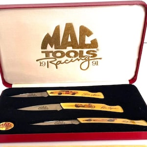 May include: A set of three folding pocket knives with yellow handles featuring racing graphics and signatures, presented in a red hinged display case. The interior lid displays the gold text MAC TOOLS 19 RACING 91, and a small gold logo emblem is included.