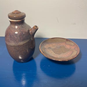 May include: A ceramic soy sauce dispenser with a matching lid and a small circular dipping dish. Both pieces feature a mottled brown and tan glaze with a smooth, glossy finish. The dispenser has a narrow spout and a rounded body shape.