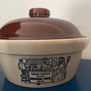 May include: A beige ceramic casserole dish with a dark brown lid. The exterior features a black ink illustration of a storefront with the text "SOLON CADDY'S FINE GROCERIES, WINES, & Co." printed on the front of the bowl, showcasing a vintage design.