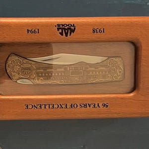 May include: A folding pocket knife with an ornate gold-tone handle featuring engraved details, displayed inside a rectangular wooden case. The case is inscribed with the text MAC TOOLS, 1938, 1994, and 56 YEARS OF EXCELLENCE on the frame.