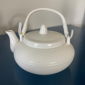 Puede incluir: Una tetera de cerámica blanca con un patrón texturizado de puntos en relieve en el cuerpo y la tapa. Incluye un pico curvo y un asa envuelta en color blanco sujeta a dos bucles metálicos verticales, rematada con un pequeño pomo puntiagudo.