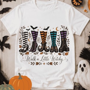 Puede incluir: Una camiseta blanca con un gráfico de cinco pares de piernas con medias a rayas y varias botas. El diseño incluye telarañas, murciélagos y hojas, con el texto "Walk a Little Witchy" impreso en letra cursiva debajo de las piernas.