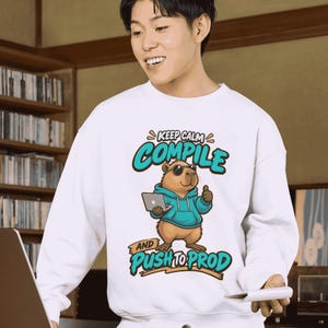 Puede incluir: Una sudadera blanca de cuello redondo con un gráfico de un capibara marrón con gafas de sol y una sudadera azul, sosteniendo un portátil y haciendo un gesto de pulgar hacia arriba. El texto dice KEEP CALM COMPILE AND PUSH TO PROD.