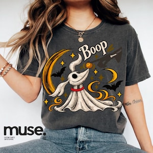 Puede incluir: Una camiseta gris carbón con un gráfico de un perro fantasma blanco con collar rojo, una luna creciente, murciélagos y estrellas. El texto Boop está impreso sobre el perro, que toca una pequeña calabaza naranja con su nariz en la escena.