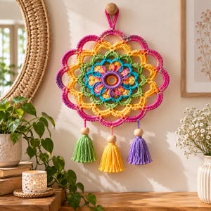 May include: A circular crochet wall hanging featuring a vibrant rainbow-coloured mandala pattern with concentric rings of pink, yellow, green, and blue yarn. Three dangling tassels in light green, yellow, and purple hang from wooden beads.