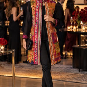 May include: A long-sleeved, knee-length jacket featuring a vibrant, multi-colored patchwork pattern with intricate embroidery and mirror accents along the front placket, worn over a solid black top and slim-fit black trousers with black heels.